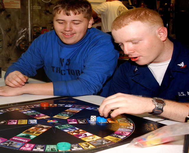 Zwei Männer spielen ein Brettspiel an einem Tisch mit Würfeln und anderen Gegenständen, während im Hintergrund ein paar Menschen stehen.