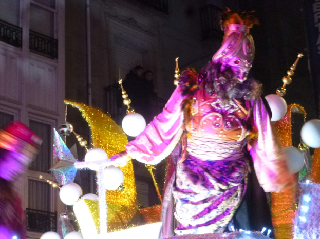 Ein Mann in einem Kostüm steht auf einem geschmückten Wagen bei einem Umzug, mit beleuchteten Gebäuden im Hintergrund.