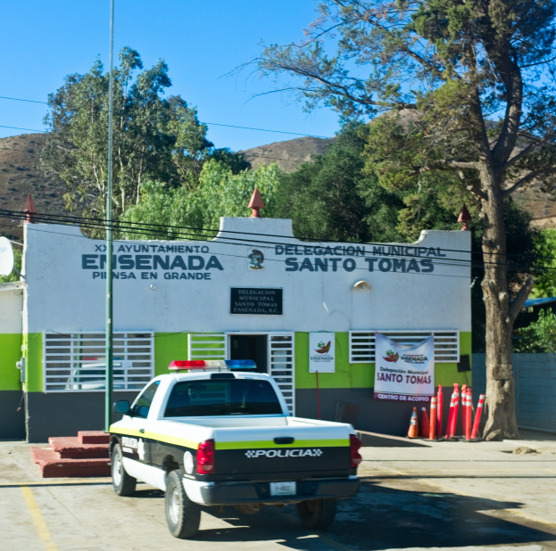 Polizeiauto vor einem Gebäude mit einem Schild, einer Fahne, Fenstern und einer Tür geparkt, mit Verkehrspfosten, einer Straßenlaterne, einer Satellitenschüssel, Kabeln, Bäumen, Hügeln und einem bewölkten Himmel im Hintergrund.
