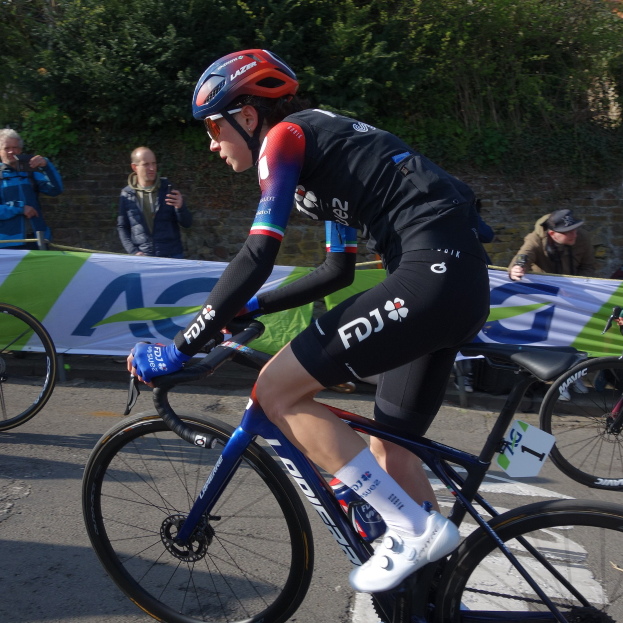 Professionelle Radsportlerin Katie Archibald mit Helm, Schutzbrille, Handschuhen und Schuhen fährt auf einem Fahrradstraße an einer Menge vorbei, mit einem Banner, einer Wand, Bäumen und einem Gebäude im Hintergrund.