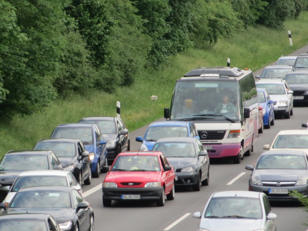 Stau auf einer Autobahn mit vielen Autos und einem Lieferwagen, Menschen in den Fahrzeugen und Bäume und Gras im Hintergrund.