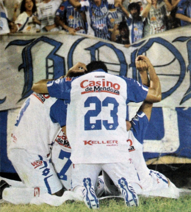 Gruppe von Fußballspielern, die ein Tor feiern, auf dem Boden mit einer jubelnden Menge im Hintergrund und einem Banner im Hintergrund.