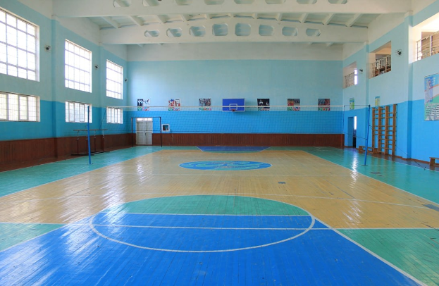 Großer Turnsaal mit einem blauen und weißen Boden, ausgestattet mit einem Basketballfeld, Netz, Pfosten, einer Tür, Fenstern, Postern an den Wänden, einem Geländer und einem Dach.