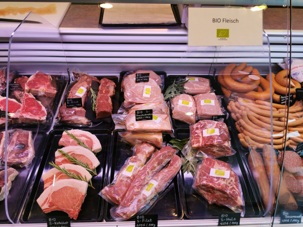 Eine Vitrine in einer Metzgerei mit verschiedenen Fleisch- und Wurstwaren in Schalen, jede mit Preisschildern, und ein Textschild im Hintergrund.