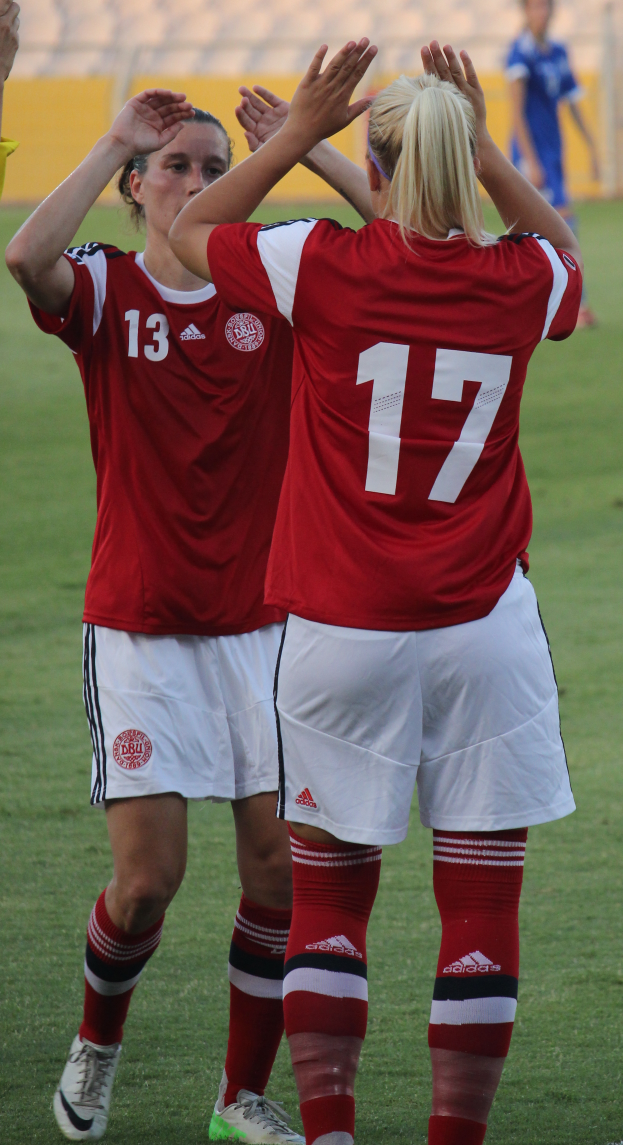Zwei Frauen in roten T-Shirts und weißen Shorts stehen auf einem Fußballfeld und klatschen begeistert, mit einer Person in einem blauen Kleid, einer gelben Tafel und ein paar Stühlen im Hintergrund.