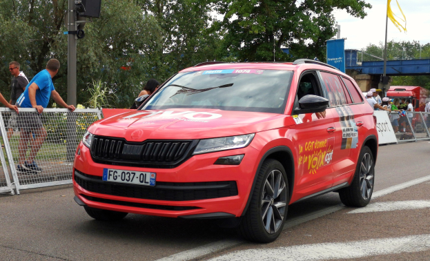 Ein rotes Auto fährt auf einer Straße neben einer Menschenmenge, einige tragen Mützen, mit Maschendrahtzaun, einer Verkehrsampelstange, Bäumen, einer Brücke, einem Fahnenmast und einem bewölkten Himmel im Hintergrund.