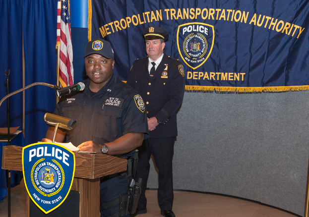 Zwei Polizeibeamte stehen vor einem Rednerpult, einer hält Papiere und spricht in ein Mikrofon; im Hintergrund sind ein Banner, eine Flagge und ein Tisch zu sehen.