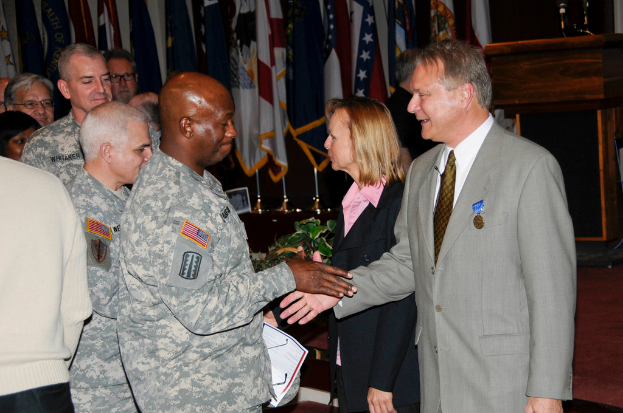 Eine Gruppe von Menschen mit zwei Personen, die sich die Hände schütteln, im Hintergrund Fahnen an Stangen, ein Podium mit einem Mikrofon und eine Topfpflanze in der Nähe.