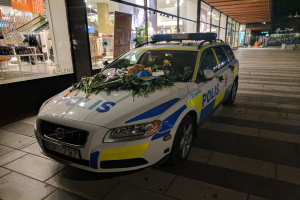 Ein mit Blumen und Blättern geschmückter Polizeiwagen parkt vor einem Geschäft mit Glaswänden und zeigt Schaufensterpuppen in Kleidern und beleuchtete Schilder im Inneren.