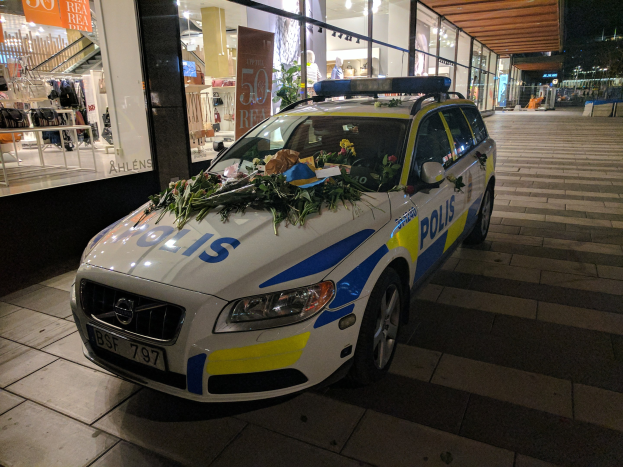 Ein mit Blumen und Blättern geschmückter Polizeiwagen parkt vor einem Geschäft mit Glaswänden und zeigt Schaufensterpuppen in Kleidern und beleuchtete Schilder im Inneren.