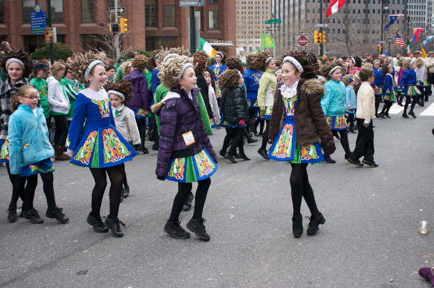 Eine Gruppe von Mädchen in Kleidern mit Jacken und Pullis tanzend auf einer Straße, mit einem roten Gebäude und einem gelben Signalmast dahinter und Gebäuden mit Flaggen an Strommasten im Hintergrund.