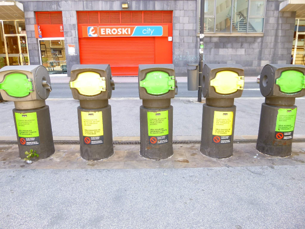 Eine Reihe von Parkautomaten entlang einer Straße, mit einem Gebäude mit Glasfenstern und einer Rolllade mit Text im Hintergrund und einem nahen Müllcontainer.