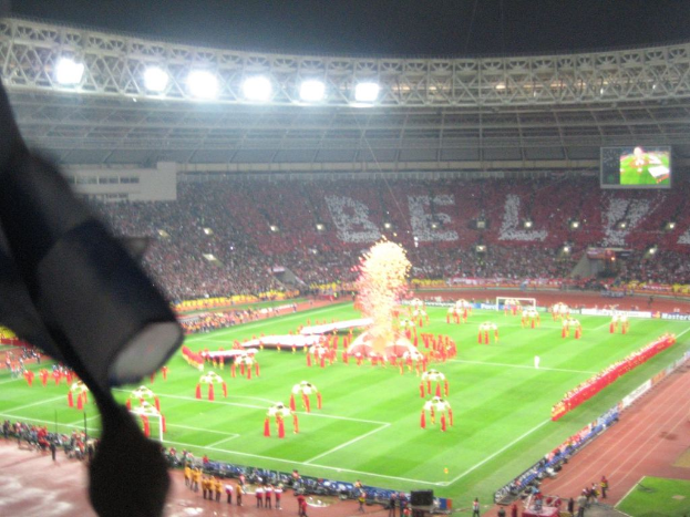 Ein Stadion mit Flutlicht, Eisengittern, Zuschauern, stehenden Individuen, Werbetafeln, Anzeigetafeln, einer Überwachungskamera und verschiedenen Flaggen und Dekorationen.
