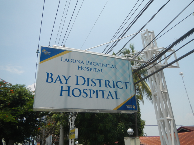 Kreiskrankenhaus Laguna mit einem Schild, Strommasten, Laternen, Bäumen, Gebäuden und einem bewölkten Himmel im Hintergrund.