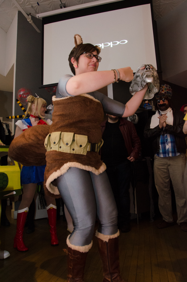 Eine Frau in einer braunen Jacke hält eine silberne Maske, während sie tanzt, und eine andere Frau in einem blauen Kleid steht und lächelt im Hintergrund, während ein weißer Projektionsschirm im Hintergrund 'Oppo' anzeigt.