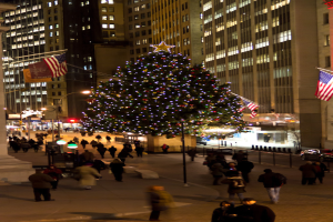 Weihnachtsbaum mit Menschen in der Mitte einer Straße, flankiert von zwei Fahnenmasten, mit beleuchteten Gebäuden im Hintergrund.