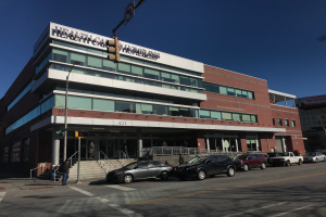 Ein großes Gebäude mit parkenden Autos davor, umgeben von Straßenlaternen, Verkehrspfosten, Verkehrssignalen, Schildern, Menschen auf dem Gehweg, Geländern, Treppen, Bäumen und einem klaren blauen Himmel, identifiziert als das New Yorker Gesundheitszentrum für Obdachlose.