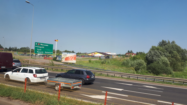 Eine Autobahn mit Autos, einem Metallzaun, Gras, Pflanzen, Bäumen, Schildern, Straßenlaternen und einem bewölkten Himmel.