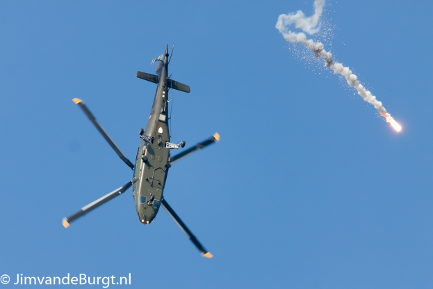 Ein militärischer Hubschrauber in der Luft, der eine Rakete hält, die Rauch und Feuer ausstößt, mit Text in der unteren linken Ecke.