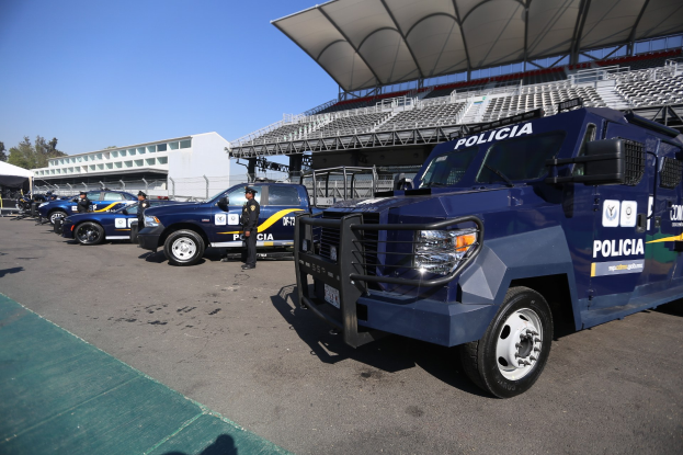 Eine Reihe von Polizeiwagen vor einem Stadion geparkt, mit Menschen im Vordergrund auf der Straße und Gebäuden, Bäumen und einem klaren blauen Himmel im Hintergrund.