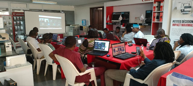 Eine Gruppe von Menschen sitzt um einen Tisch mit Laptops, Mäusen, Flaschen und anderen Gegenständen und nimmt an einem digitalen Marketing-Workshop teil, mit einem Banner, einem Display, einer Tür, Deckenleuchten und elektronischen Geräten im Hintergrund.