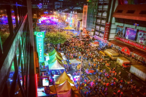 Eine bunte Faschingszene mit einer großen Menschenmenge auf der Straße, umgeben von Zelten, Geschäften, Gebäuden und Bannern, mit zahlreichen Lichtern, die den Hintergrundbereich erhellen.