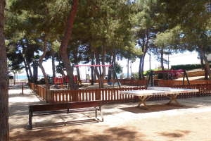 Eine Parkszene mit einem zentralen Tischtennistisch, umgeben von Bänken, einem Zaun, Schaukeln, Pfählen, Pflanzen und Bäumen, mit einem Wasserlauf und einem klaren blauen Himmel im Hintergrund.