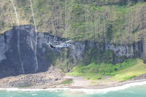 Ein Hubschrauber fliegt über das Meer in der Nähe einer Klippe, mit Wasser darunter, Felsen und Gras am Boden und Bäumen auf dem Berg im Hintergrund.