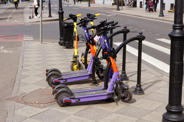 Eine Reihe geparkter E-Scooter auf einer Straße mit Fußgängern, Radfahrern, einem Müllcontainer und Gebäuden im Hintergrund.