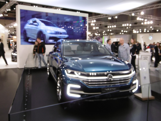 Ein blauer Volkswagen Touareg wird auf der IAA in Frankfurt präsentiert, umgeben von Menschen in verschiedenen farbigen Kleidern, mit einem großen Bildschirm und Deckenleuchten im Hintergrund.