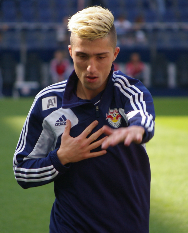 Ein junger Mann in einem blauen T-Shirt steht auf einem Fußballfeld und streckt die Arme aus, wobei er entschlossen wie ein Flügelstürmer wirkt.