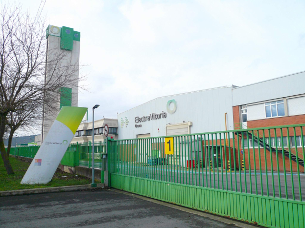 Ein großes Gebäude mit einem grünen Zaun davor, umgeben von einer Straße, Gras, Bäumen, Laternenmasten und einer Tafel, unter einem bewölkten Himmel, identifiziert als Hauptsitz von Electrovictoria.