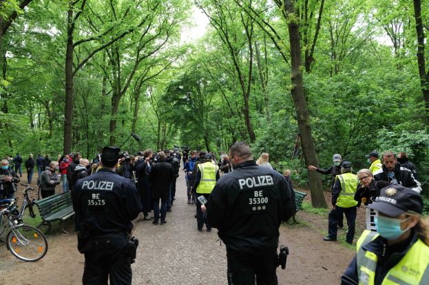 Eine Gruppe von Polizisten vor einer Menge bei einer Anti-Terror-Demonstration in Berlin, mit Fahrrädern und einer Bank im Vordergrund und Bäumen im Hintergrund.
