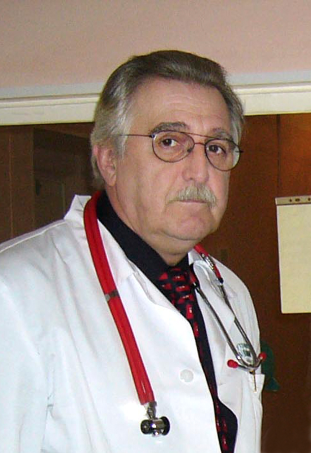 Ein Mann in einem weißen Kittel mit einem Stethoskop um den Hals steht vor einer Wand mit einem daran befestigten Blatt Papier und sieht ernst aus.