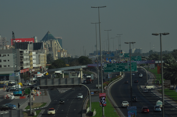 Eine Luftaufnahme einer Stadt mit Geb├Ąuden, T├╝rmen, Kraftfahrzeugen, Werbetafeln, Schildern, Stra├čen, B├Ąumen, Stra├čenlaternen, Menschen, Verkehrskegeln und Informationsschildern unter einem klaren Himmel.