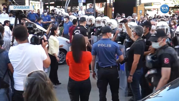 Eine Gruppe von Menschen vor einer Reihe von Helmträgern der Polizei mit Kameras, mit Fahrzeugen, Gebäuden und Schildern, darunter eines mit der Aufschrift "türkische Polizei bei Zusammenstöüen mit Demonstranten in Ankara" im Hintergrund.