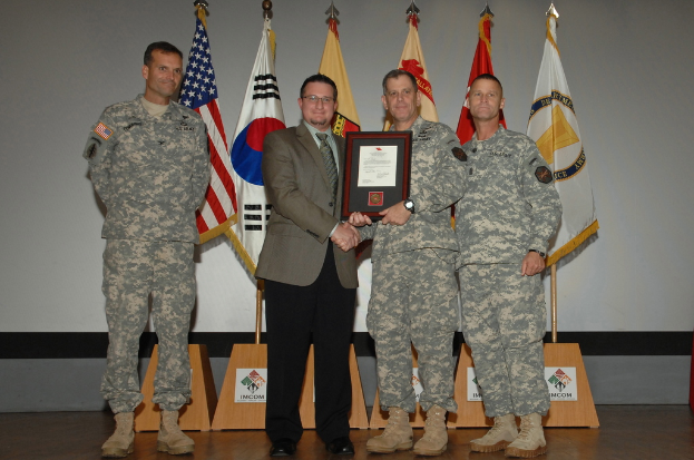 Vier Männer stehen auf einer Bühne: drei in Armeeuniformen und einer in Blazer, Krawatte und Hemd, die Hände schütteln und einen Preis überreichen, mit sechs Fahnen an Stangen und einer weißen Wand im Hintergrund.