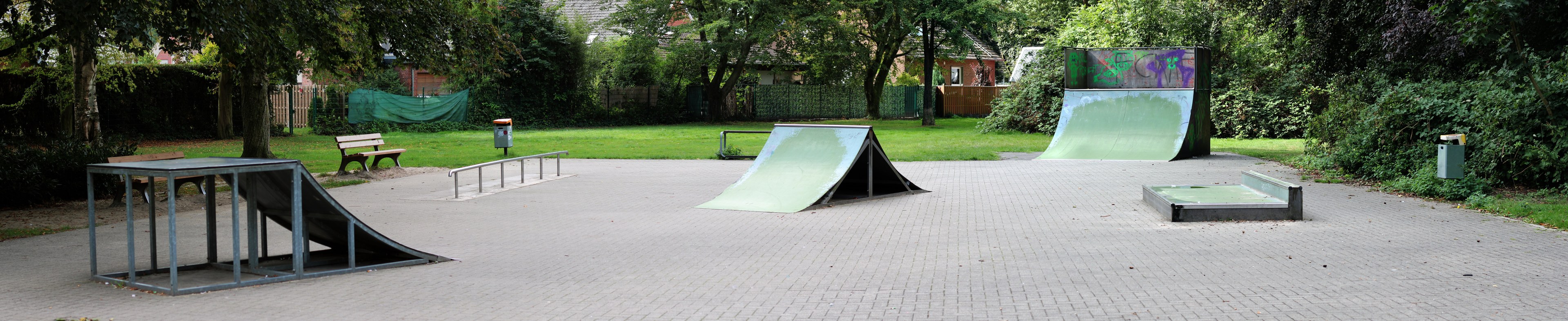 Ein Park mit einer zentralen Skaterrampe, umgeben von Bänken, Geländern, Gras, Pflanzen, Bäumen und Gebäuden im Hintergrund.