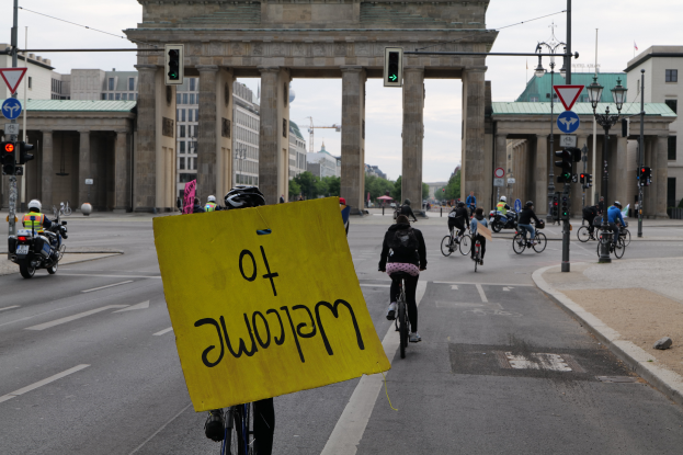 Eine Gruppe von Radfahrern mit Helmen fährt eine Straße entlang vor dem Brandenburger Tor in Berlin, Deutschland, wobei eine Person ein gelbes Schild hält, Lichtmasten, Verkehrszeichen, Gebäude, Bäume und ein klarer blauer Himmel im Hintergrund zu sehen sind.