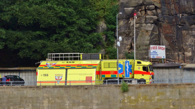 Ambulanz am Straßenrand neben einer Klippe geparkt, mit verschiedenen Gegenständen im Vordergrund und Bäumen und einem Auto im Hintergrund.