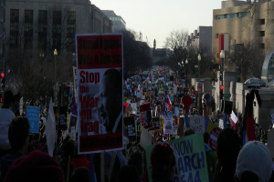 Menschen protestieren auf einer Straße mit Plakaten, mit Gebäuden, Bäumen, Laternen und dem Himmel im Hintergrund.