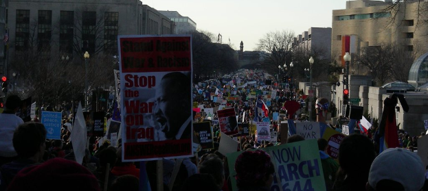 Menschen protestieren auf einer Straße mit Plakaten, mit Gebäuden, Bäumen, Laternen und dem Himmel im Hintergrund.