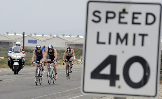 Gruppe von Menschen mit Helmen auf Fahrrädern auf einer Straße neben einem Tempolimit-Schild, mit Gebäuden, Pfählen, Pflanzen, Bäumen und einem klaren blauen Himmel im Hintergrund.