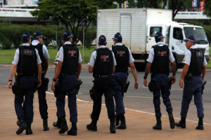Eine Gruppe von Beamten in Uniform, einschließlich Mützen, Hemden, Schutzwesten, Uhren, Hosen und Schuhen, geht auf einer Straße. Rechts daneben parkt ein Lastwagen in der Nähe eines Zauns, mit Gebäuden, Bäumen, Pflanzen, Polen und Bannern im Hintergrund. Eine Brücke ist oben rechts zu sehen.