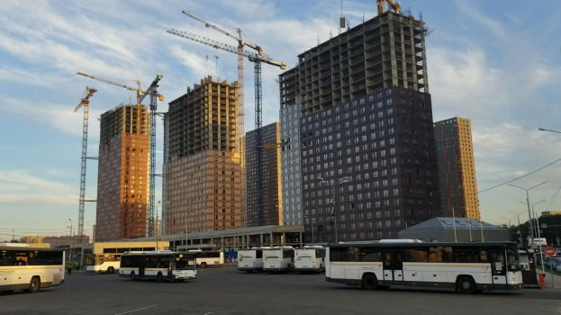 Eine Gruppe von Bussen, die auf einem Parkplatz neben hohen Gebäuden geparkt sind, mit Straßeninfrastruktur und Bäumen unter einem bewölktem Himmel.