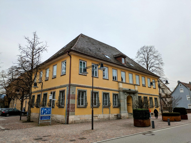 Ein gelbes Eckgebäude, das ehemalige Rathaus in Heidelberg, Deutschland, mit Straßenlaternen, Schildern, Fahrzeugen, Menschen, Büschen, Bäumen und einem klaren blauen Himmel.