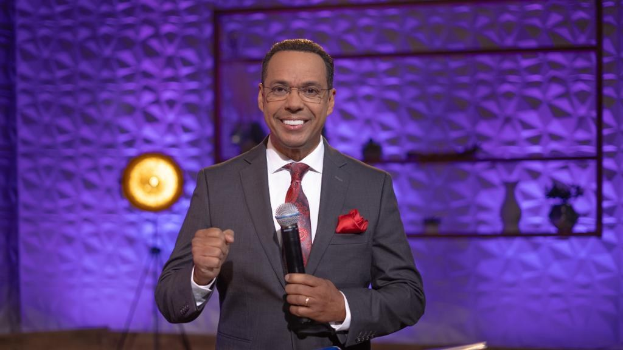 Stephen Colbert, Moderator von "The Daily Show with Trevor Noah", steht auf einer Bühne und hält ein Mikrofon in der Hand, trägt einen Anzug, eine Krawatte und eine Brille, mit einer Wandhalterung und einer Lampe dahinter.