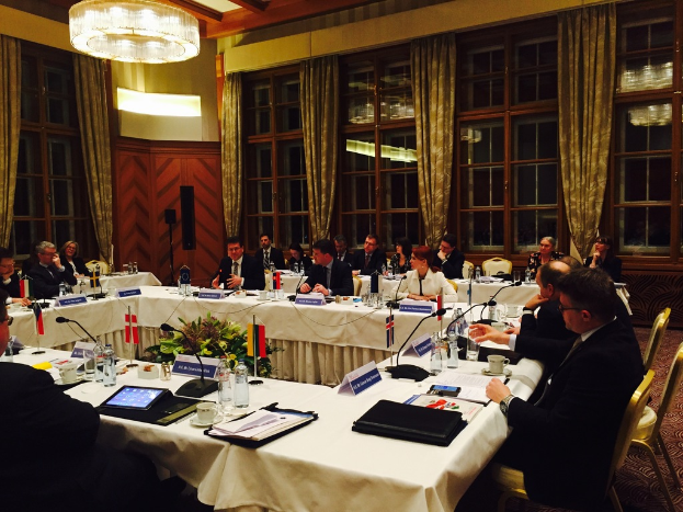 Menschen an einem Tisch mit verschiedenen Büromaterialien, darunter Ordner, Papiere, Getränke und elektronische Geräte, in einem Konferenzraum mit roter Wand, grünen Vorhängen und einer sichtbaren Decke.