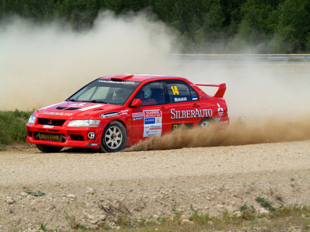 Ein roter Rallye-Wagen mit Text und Nummern fährt auf einer von Steinen und Gras bedeckten Schotterstraße, mit zwei Helmträgern im Inneren, während Rauch aufsteigt und Bäume im Hintergrund stehen.
