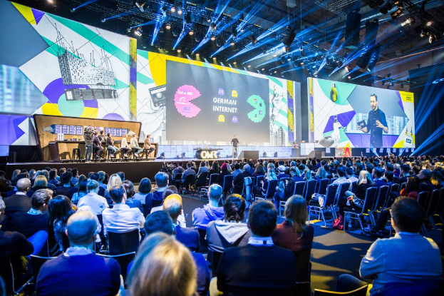 Eine große Menschenmenge, die auf Stühlen vor einer Bühne sitzt, mit einem Tisch voller Gegenstände links und Bildschirmen und Lichtern im Hintergrund.